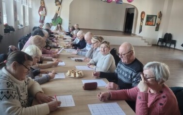Seniorzy Klubu Seniora w Nożynie siedzą przy wsp&oacute;lnym stole, mając przed sobą plansze i grają w grę ,,Bingo&rsquo;&rsquo;