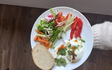 Na talerzu znajduje się posiłek o nazwie ,,Jajko po turecku&rdquo; składający się z  pokrojonych warzyw tj. marchew, sałata, og&oacute;rek świeży, seler naciowy, rzodkiewka, pomidorki, papryka oraz pieczywa i sadzonego jajka podanego na sosie.