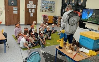Grupa dzieci z OWP w Czarnej Dąbr&oacute;wce siedzi w klasie na dywanie przed nimi stoi pszczelarz prezentujący sw&oacute;j zaw&oacute;d.  