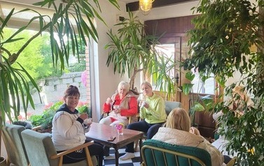 Kobiety siedzą wok&oacute;ł stolika w kawiarni i jedzą desery z puchark&oacute;w. Otaczają je wysokie rośliny doniczkowe.