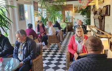 Osoby siedzą przy stolikach w kawiarni z dużymi roślinami, wiklinowymi fotelami i podłogą w czarno-białą szachownicę.
