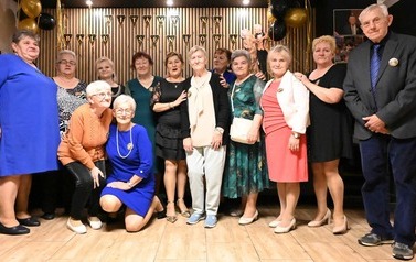 Uczestnicy Balu Seniora pozują do wsp&oacute;lnego zdjęcia na tle powitalnego napisu i czarno-złotych balon&oacute;w. Większość stoi blisko siebie, dwie kobiety pozują niżej z przodu.