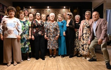 Grupa elegancko ubranych kobiet i mężczyzna w marynarce pozują do wsp&oacute;lnego zdjęcia. W tle złote i czarne balony oraz ozdobna girlanda.