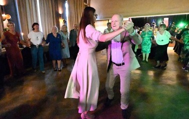 Mężczyzna w jasnej marynarce tańczy w parze z kobietą w długiej, jasnej sukience. Wok&oacute;ł nich inni uczestnicy trzymają się za ręce.