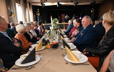 Goście Balu Seniora siedzą po obu stronach długiego stołu udekorowanego kwiatami, świecami oraz złotymi i czarnymi serwetkami. W tle na scenie występuje zesp&oacute;ł muzyczny.
