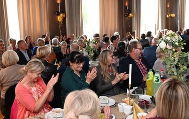 Uczestnicy Balu Seniora siedzą przy zastawionych stołach i klaszczą. Salę zdobią kompozycje kwiatowe oraz czarne i złote balony.