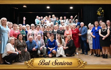 Uczestnicy Balu Seniora pozują do wsp&oacute;lnego zdjęcia przed sceną. Fotografię otacza ozdobna złota ramka z napisem &bdquo;Bal Seniora&rdquo;.
