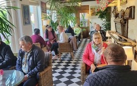 Osoby siedzą przy stolikach w kawiarni z dużymi roślinami, wiklinowymi fotelami i podłogą w czarno-białą szachownicę.