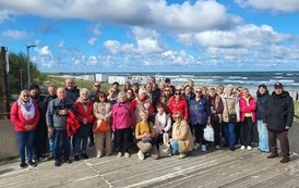 Grupa os&oacute;b pozuje na drewnianym tarasie nad morzem. W tle widać plażę, wzburzone fale i chmury.
