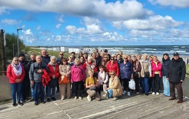 Grupa os&oacute;b pozuje na drewnianym tarasie nad morzem. W tle widać plażę, wzburzone fale i chmury.